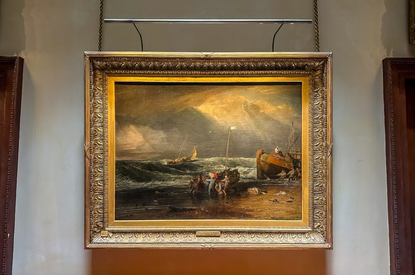 A framed painting in the style of Turner Art depicts a stormy seascape with crashing waves, boats near the shore, and figures on the beach under a cloudy sky, hanging on a gallery wall.