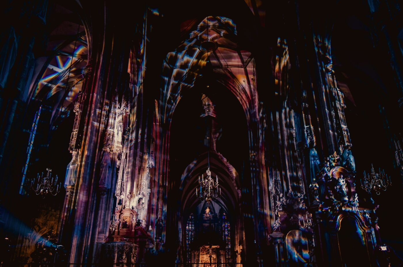 Dimly lit cathedral interior with tall arches and ornate columns. Wicked multicolored light patterns are projected onto the walls and ceiling, creating a dramatic and mystical atmosphere within the gothic architecture.