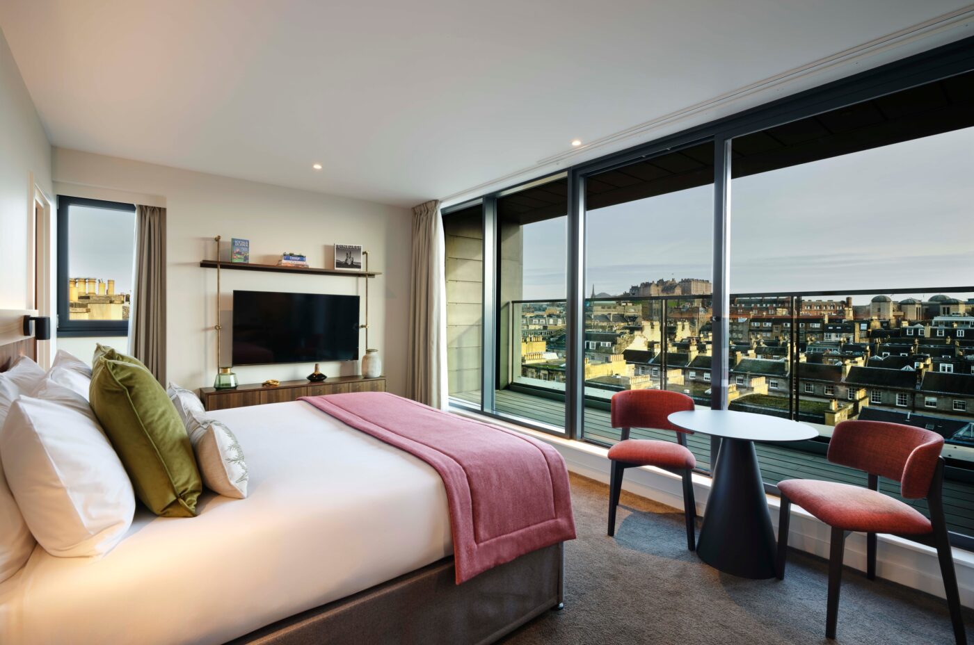 Modern hotel room with a large bed, colorful pillows and blanket, TV, two red chairs, a small round table, and floor-to-ceiling windows overlooking a cityscape with buildings under a cloudy sky.