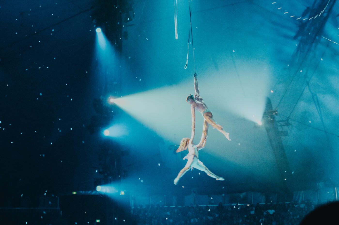 Two aerial acrobats perform high above a crowd, suspended by ropes under blue stage lights in a large, darkened arena. The performers are mid-air, holding onto each other with dramatic lighting behind them.