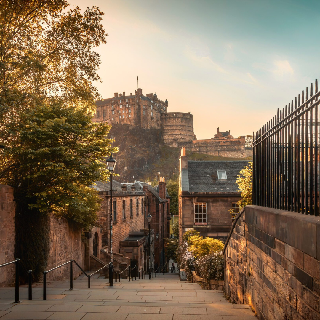 A stone stairway lined with railings leads up to Edinburgh Castle, which sits atop a rocky hill under a soft, golden sky; historic buildings and trees frame the scene.
