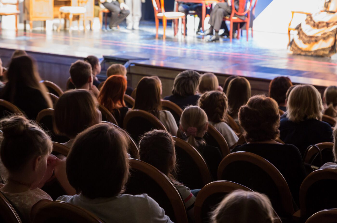An audience sits in rows of seats watching a live stage performance of Wicked. The focus is on the backs of the audience, with the brightly lit stage and actors visible in the background.