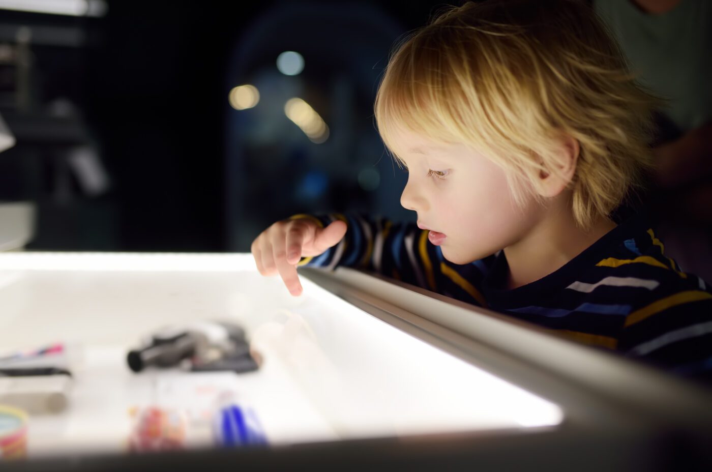 A young child with blond hair and a striped shirt looks intently at objects displayed in a well-lit glass case, curiosity sparked by the wicked allure of mysterious treasures against a dark, blurred background.