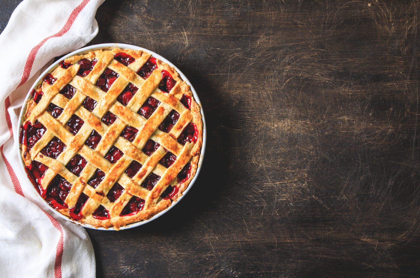 A freshly baked cherry pie with a golden lattice crust sits on a dark wooden surface, partially wrapped in a white cloth with red stripes, as if waiting to be enjoyed on a special One Day.