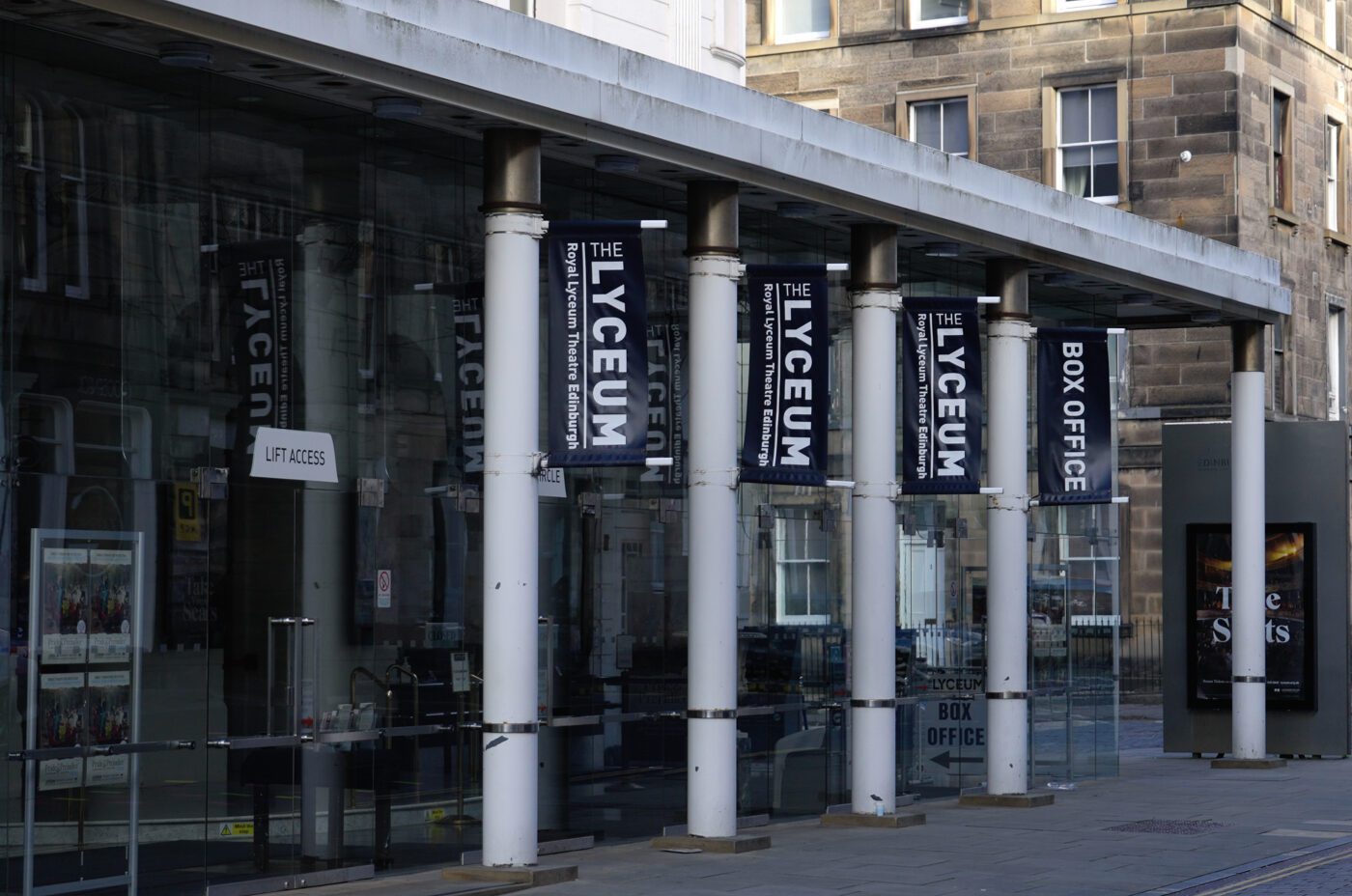 The entrance of The Lyceum theatre—with glass walls, white pillars, black and white signs, and a box office—welcomes visitors during the International Children's Festival. The street and a few parked cars can be seen in the background.