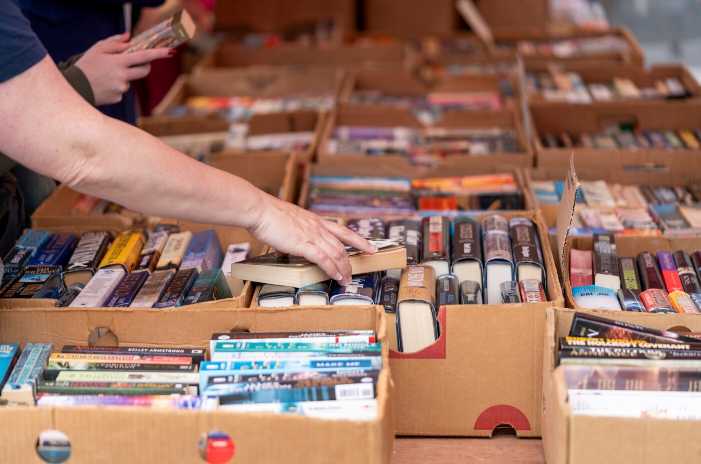 A person reaches for a book among rows of books neatly arranged in cardboard boxes at a biennial outdoor book market or sale.